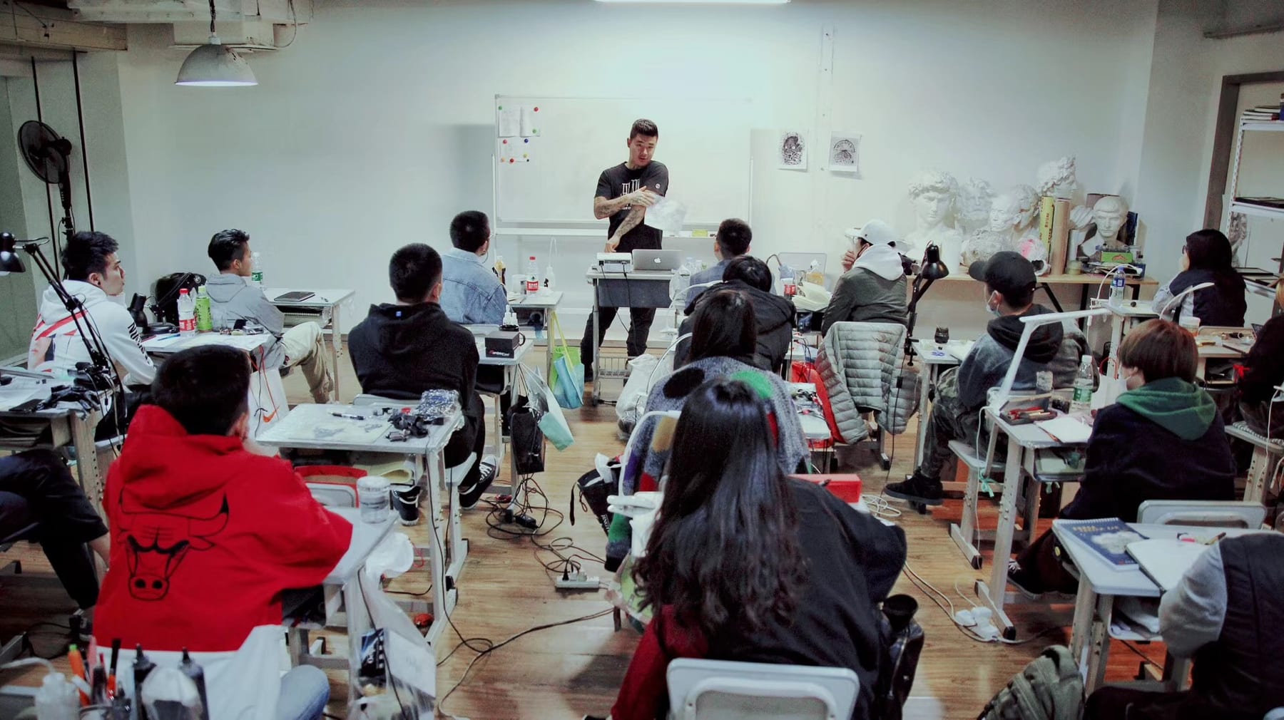 Wang Lei leading a small-group realism tattoo mentorship class at the Lei Shi Beijing headquarters — students at workstations with practice skins, classical plaster busts on the back shelf
